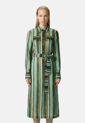 Vrouw draagt een groenbruine verticale gestreepte overhemdjurk met lange mouwen, knopen, borstzakjes en een strikceintuur in de taille.