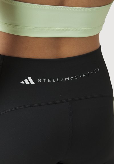 Leggings sportivi neri con una cintura larga che presenta il logo "STELLAMCCARTNEY" e dettagli testurizzati. Un reggiseno sportivo di un verde chiaro è sopra.