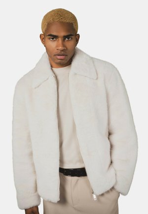 Veste en fausse fourrure blanche avec un col chemise et une fermeture à glissière. Texture douce, coupe décontractée, et poches latérales. Portée sur un haut beige.