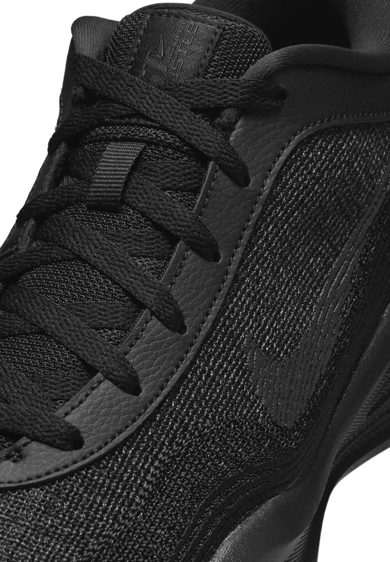 Zapatilla deportiva negra con parte superior sintética texturizada, cordones planos negros y un diseño elegante con un sutil acento de logo en el lateral.