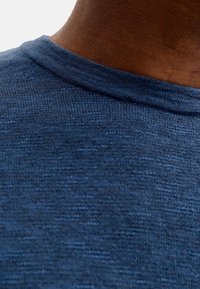 Gros plan sur le cou et l'épaule supérieure d'une personne à la peau foncée portant un pull tricoté bleu texturé sur un fond uni.