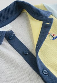 Detailný záber na detskú polokošeľu s tmavomodrým golierom a gombíkmi, sivým predným dielom a žltým vnútorným lemovaním s malou výšivkou plachetnice.