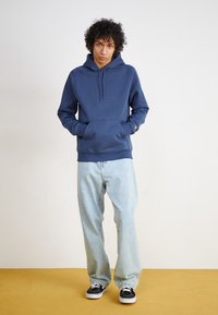 Navy blue hoodie with a front pocket and drawstring hood, paired with light denim jeans and black sneakers. Smooth fabric texture.
