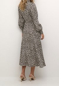 Frau in einem langärmeligen, knöchellangen Kleid mit Leopardenmuster, die beige High-Heel Slingback-Schuhe trägt und vor einem einfarbigen Hintergrund steht.