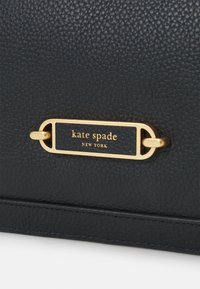 Czarna torebka ze skóry o teksturowanej powierzchni, z prostokątną złotą tabliczką z logo z napisem „kate spade NEW YORK” w czarnych literach.