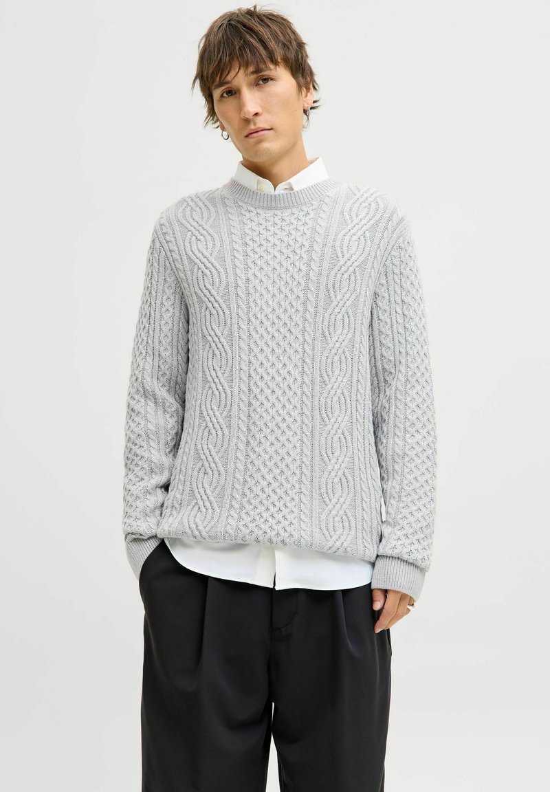 Hellgrauer Strickpullover mit Kabelmustern, gerippten Bündchen und hohem Kragen, getragen über einem weißen Hemd mit Kragen, kombiniert mit dunklen Hosen.