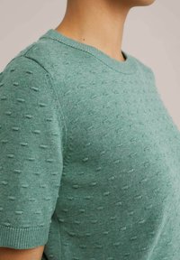 Lichtgroene gebreide trui met een zachte textuur, korte mouwen en een ribbelboord, versierd met kleine gepofte stippenpatronen.