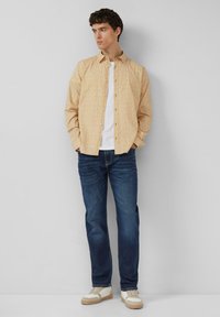 Gele overhemd met patroon en knoopsluiting, gecombineerd met donkerblauwe rechtvallende jeans en witte schoenen met beige accenten.