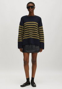Oversized Pullover in Marineblau und Olivgrün gestreift mit weichem Material, kombiniert mit einem schwarzen Lederminirock und flachen Schuhen.