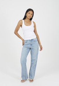 Kobieta z długimi ciemnymi włosami, ubrana w białą koszulkę na ramiączkach, jasnoniebieskie szerokie dżinsy, srebrne kolczyki w kształcie obręczy i białe sandały na obcasie, stojąca na białym tle.