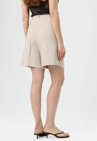 Shorts beige arrivant aux genoux, coupe ajustée avec plis, taille haute et poches latérales. Texture lisse et esthétique minimaliste.