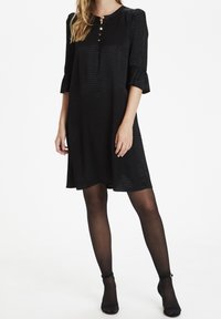 Svart skjortklänning med strukturerad tyg, rund halsringning, tre-knapp framsidan och klockärmar. Stylerad med svarta strumpbyxor och klackar.