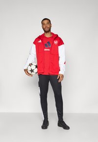 Red and white zip-up jacket over a red t-shirt with a black logo, black athletic pants with white stripes, and black shoes, holding a soccer ball.