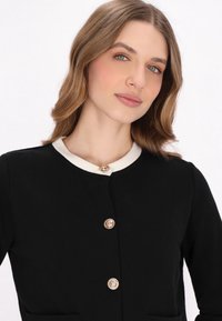 Jachetă din material textil negru, cu guler alb, detalii cu nasturi aurii și buzunare cu desfăcut, având un design structurat și o textură netedă.