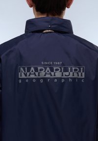 Mørkeblå vandtæt jakke med høj krave, udstyret med et "NAPAPIJRI" logo og teksten "SINCE 1987" på ryggen i kontrasterende lys grå.