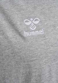 Camiseta de algodón gris con un logo de abeja bordado en blanco y el texto "hummel" en una fuente deportiva en el área del pecho. Tejido suave y texturizado.