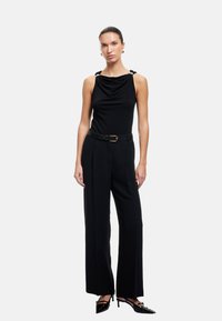 Top negro con drapeado y tirantes acentuados con nudos, combinado con pantalones de pierna ancha de talle alto y zapatos de tacón de punta. Textura de tela suave.
