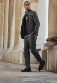 Gray tweed suit with a tailored fit, white shirt, and blue paisley scarf. Black shoes with brown soles and visible lacing details.