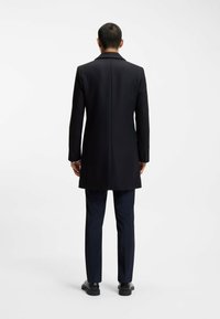 Cappotto in misto lana nero con vestibilità sartoriale, maniche lunghe e colletto a punte, abbinato a pantaloni blu navy e scarpe nere eleganti.
