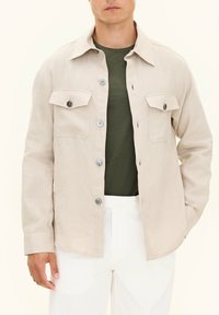 Beige skjorte med knapper lavet af letvægtsstof. Har to brystlommer, matchende knapper og en afslappet pasform. Bæres over en grøn T-shirt.