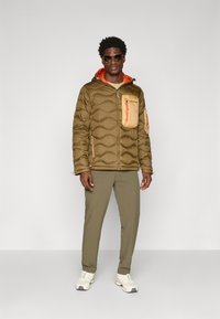 Bruine pufferjack met een opvallende oranje capuchon en borstzak, gecombineerd met olijfgroene broek en witte sneakers.