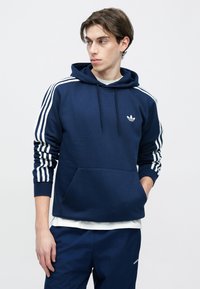 Donkerblauwe capuchontrui met witte strepen op de mouwen, een voorzak en een klein logo. Gemaakt van zacht materiaal met een losse pasvorm.