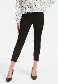 Kvinna som bär svarta korta smala byxor med ankelblixtlås, en vit blus med svarta prickar och svarta spetsiga klackskor.