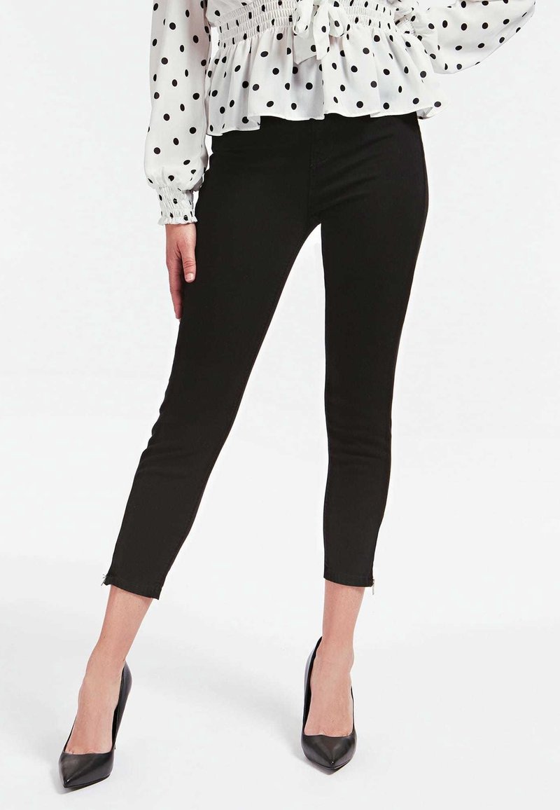 Kvinna som bär svarta korta smala byxor med ankelblixtlås, en vit blus med svarta prickar och svarta spetsiga klackskor.