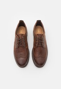 Chaussures en cuir marron avec un bout arrondi, un design de type wingtip et des accents perforés. Elles présentent des œillets gris et des lacets marron.