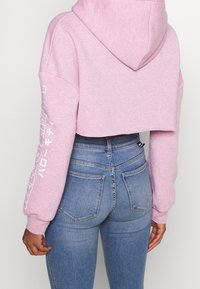 Osoba v růžové krátké mikině s bílým nápisem na rukávu a modrých rifleách s vysokým pasem, zobrazená zezadu na jednobarevném pozadí.