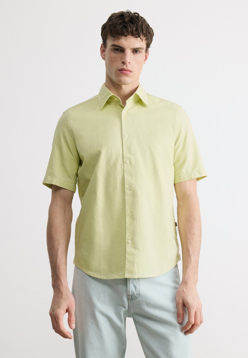 Jeune homme portant une chemise boutonnée à manches courtes jaune clair et un jean bleu clair, debout devant un fond blanc uni.