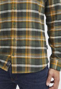 Grøn, gul og hvid ternet flannel skjorte med knaplukning foran og grønne knapper, udstyret med en buet kant og mærkedetaljer på siden.