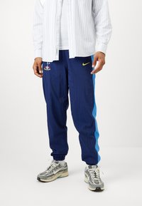 Marineblaue Jogginghose mit einem blauen Seitenstreifen, ausgestattet mit einem bunten bestickten Design und einem gelben Nike-Logo in der Nähe des Taillenbundes.
