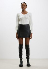 Cardigan grigio chiaro lavorato a maglia con design testurizzato e bottoni, abbinato a una mini gonna di pelle nera e stivali neri alti fino al ginocchio.