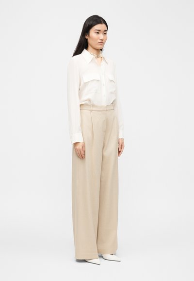 Γυναίκα που στέκεται στραμμένη προς τα αριστερά, φορώντας λευκό πουκάμισο με κουμπιά, μπεζ παντελόνι φαρδιάς γραμμής, λευκά μυτερά παπούτσια και περιδέραιο με μαργαριτάρια.