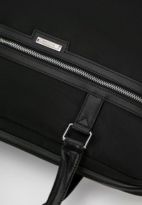 Zwarte textiel tas met een soepel lederen accent, horizontale ritsafsluiting en metalen hardware. Bevat een label met de merknaam.