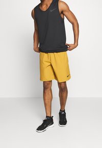 Débardeur noir Nike, short jaune avec logo noir, et chaussures de sport noires, debout sur un fond neutre. Design minimaliste, matériaux légers.