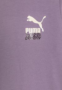 Vijolična majica iz mehkega materiala s belim logotipom Puma in grafičnim besedilom spodaj z valovitim dizajnom v črni in sivi barvi.