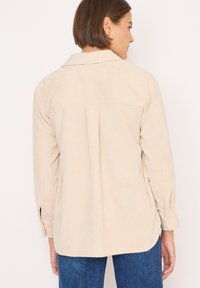 Camicia in corduroy beige con colletto, maniche lunghe e dettagli con bottoni. Caratterizzata da una texture verticale e una vestibilità rilassata con una leggera coda.