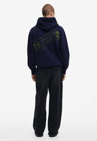 Marineblauwe hoodie met een trekkoordcapuchon, voorzien van een gestructureerde grafiek in limoengroen op de achterkant; gecombineerd met losse zwarte broek.