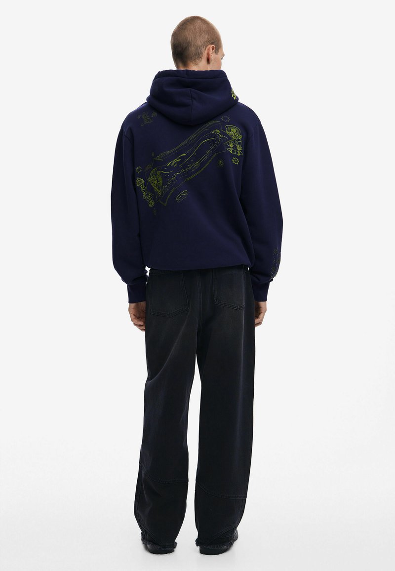 Marineblauwe hoodie met een trekkoordcapuchon, voorzien van een gestructureerde grafiek in limoengroen op de achterkant; gecombineerd met losse zwarte broek.