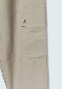 Tissu beige avec une poche rectangulaire dotée d'un seul bouton beige et des coutures visibles le long des bords.