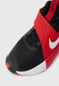 Chaussure de basketball Nike noire, rouge et blanche avec un bout texturé, une large sangle rouge sur le milieu du pied et un logo swoosh blanc sur le côté.