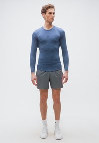 Άνδρας που στέκεται, φοράει μπλε αθλητικό πουκάμισο με μακριά μανίκια, γκρι σορτς, λευκές κάλτσες Nike και λευκά αθλητικά παπούτσια, μπροστά σε απλό ανοιχτό φόντο.