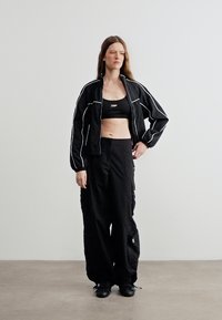 Femme en veste de sport noire avec des passepoils blancs, brassière de sport noire, pantalon cargo noir ample et baskets noires, debout contre un mur clair uni.