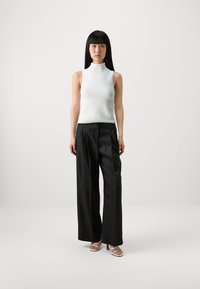 White sleeveless top with a high neck, paired with black wide-leg trousers. The model wears beige strappy heels, standing against a plain background.