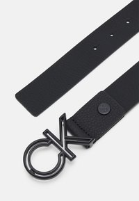 Cintura in pelle nera con finitura testurizzata, dotata di una grande fibbia in metallo nero a forma di "CK." Cinghia piatta con diversi fori per la regolazione.