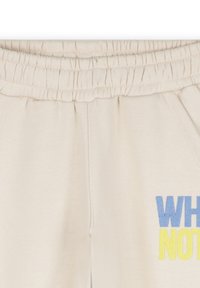 Crème kleurige sweatpants met een elastische tailleband, voorzien van blauwe en gele tekst "WHY NOT" aan de voorkant. Zachte katoen textuur, ontspannen pasvorm.