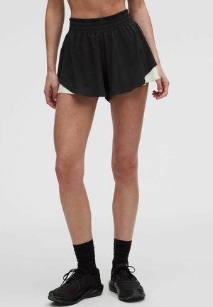 Kvindelig model iført sorte atletikshorts med hvid inderside, sorte sokker og sorte løbesko mod en ensartet lys baggrund.