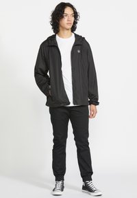 Sort letvægtsjakke med hætte, lynlås foran, elastiske manchetter og sidelommer, båret over en hvid T-shirt og sorte bukser med sneakers.